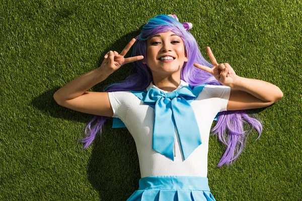 Sorrindo asiático anime menina deitado na grama e mostrando sinais de paz — Fotografia de Stock