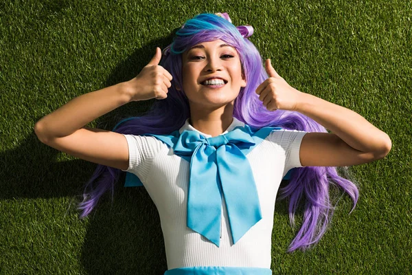 Smiling asian anime girl lying on grass and showing thumbs up — Stock Photo