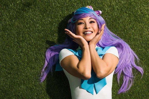 Sonriente asiático anime chica en púrpura peluca mintiendo en verde hierba - foto de stock