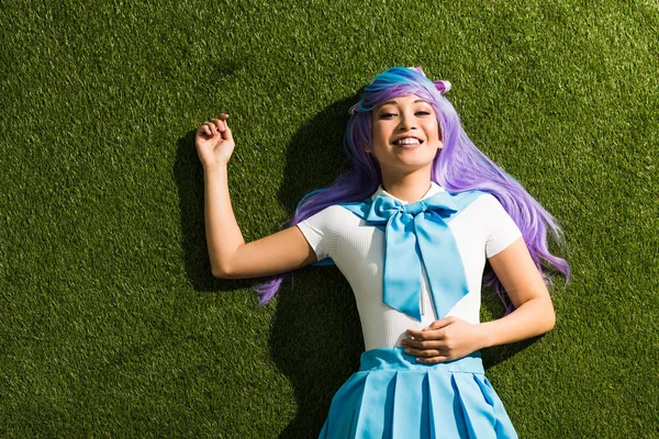 Sonriente asiático anime chica en púrpura peluca mintiendo en verde hierba - foto de stock