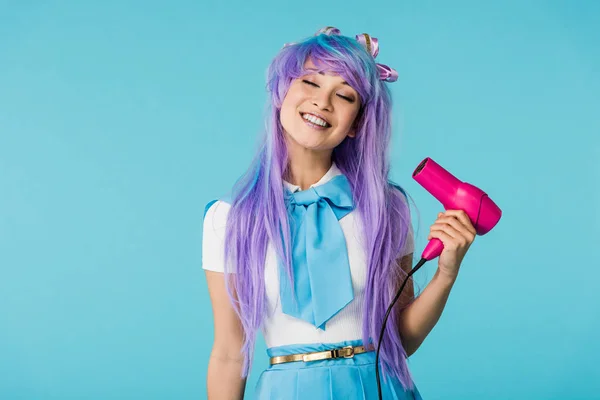 Sorrindo anime menina na peruca usando secador de cabelo com olhos fechados isolado em azul — Fotografia de Stock