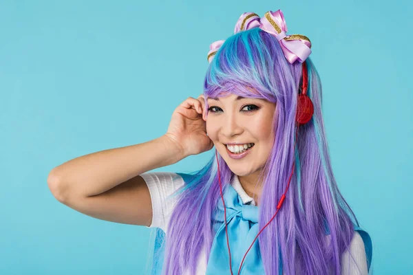 Улыбающаяся аниме девушка в фиолетовом парике слушает музыку в наушниках, изолированных на синем фоне — стоковое фото