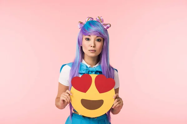 Chica anime en peluca púrpura sosteniendo encaprichamiento emoticono aislado en rosa - foto de stock