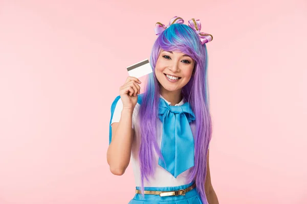 Улыбающаяся азиатская девушка аниме в фиолетовом парике с кредиткой, изолированной на розовом — стоковое фото
