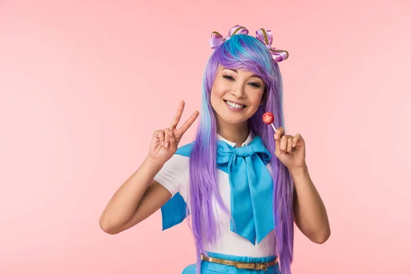 Happy asian anime girl holding lollipop and showing peace sign isolated on pink — Stock Photo