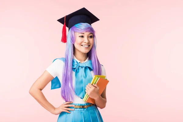 Smiling asian anime girl in academic cap holding books isolated on pink — Stock Photo