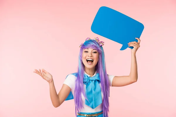 Возбужденная азиатская девушка аниме в фиолетовом парике, держащая синий пузырь речи на розовом — стоковое фото
