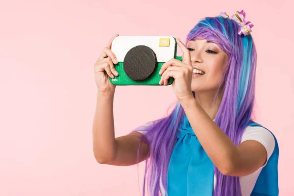 Smiling asian anime girl in purple wig holding cardboard camera isolated on pink — Stock Photo