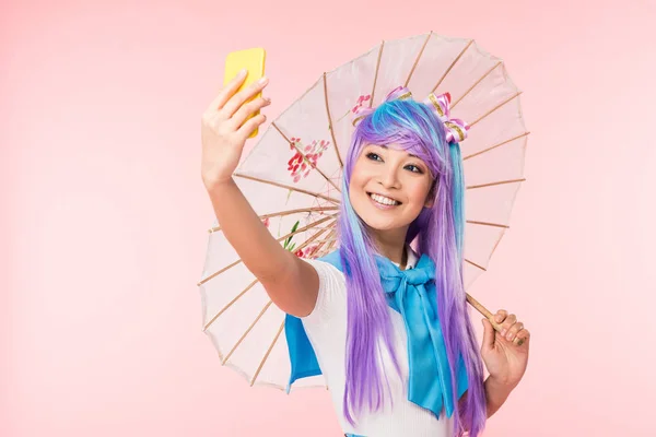 Smiling asian anime girl with paper umbrella taking selfie isolated on pink — Stock Photo