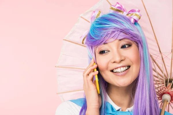 Smiling asian anime girl holding paper umbrella and talking on smartphone isolated on pink — Stock Photo