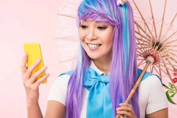 Sourire asiatique anime fille avec papier parapluie regarder smartphone isolé sur rose — Photo de stock