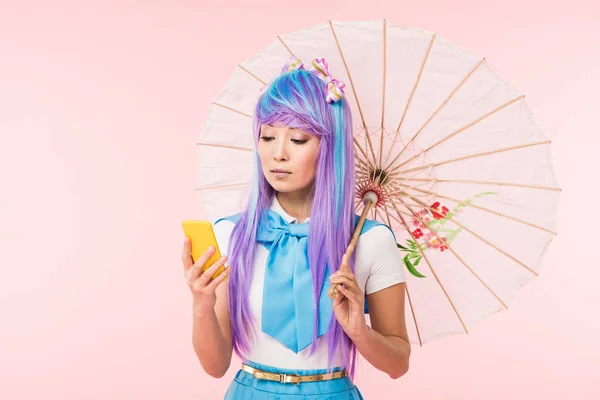 Anime asiático niña sosteniendo paraguas de papel y el uso de teléfono inteligente aislado en rosa - foto de stock