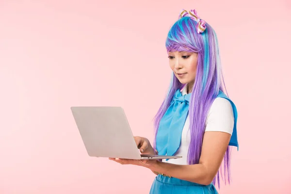 Hermosa chica de anime en peluca púrpura usando portátil aislado en rosa - foto de stock