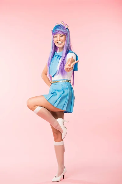 Largura completa vista de feliz asiático anime chica de pie en una pierna y mostrando la paz signo en rosa - foto de stock