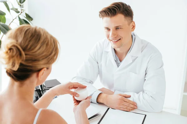 Vue arrière de la femme tenant récipient avec crème cosmétique près médecin heureux — Photo de stock