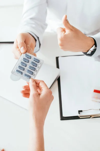 Cropped view of doctor in white coat showing thumb up while giving pills to patient — Stock Photo