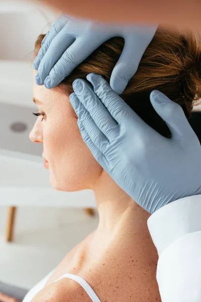 Vista recortada del dermatólogo en guantes de látex examinando el pelo del paciente en la clínica - foto de stock