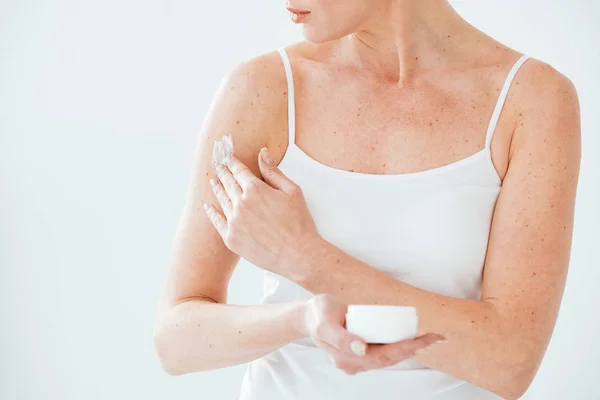 Cropped view of woman holding container and applying cosmetic cream on white — Stock Photo