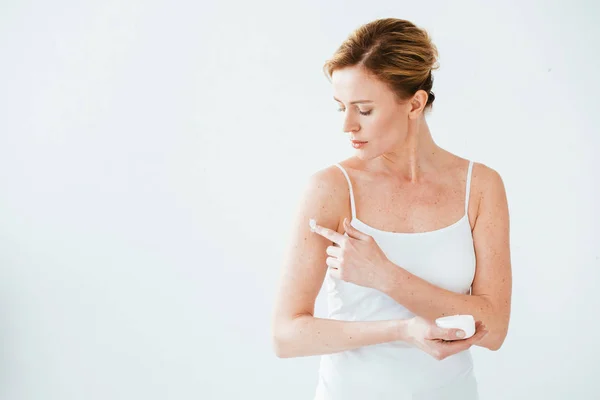 Mulher atraente com doença de pele segurando recipiente e aplicando creme cosmético no branco — Fotografia de Stock