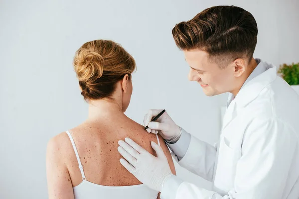 Glücklicher Dermatologe in Latex-Handschuhen mit Filzstift in der Nähe einer Frau mit Melanom isoliert auf weiß — Stockfoto