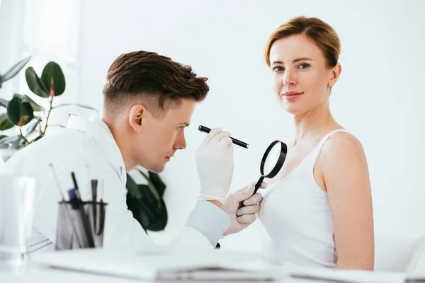 Guapo dermatólogo sosteniendo lupa mientras examina atractiva mujer con melanoma - foto de stock