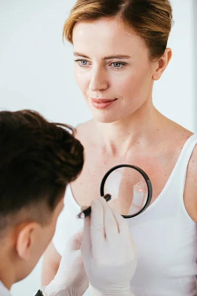 Cropped view of dermatologist holding magnifying glass while examining attractive woman with melanoma — Stock Photo