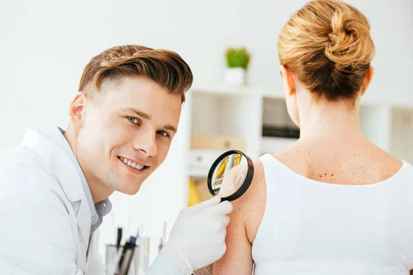 Cheerful dermatologist holding magnifying glass while examining woman with melanoma — Stock Photo