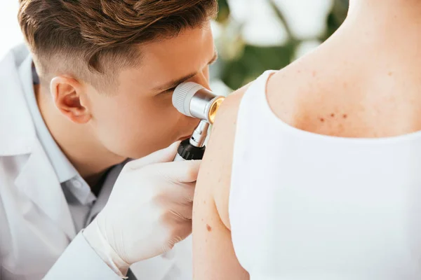 Dermatologista em luvas de látex segurando dermatoscópio enquanto examina paciente com doença de pele — Fotografia de Stock
