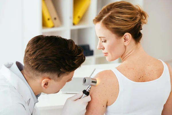 Dermatologista em luvas de látex segurando dermatoscópio enquanto examina paciente atraente com doença de pele — Fotografia de Stock