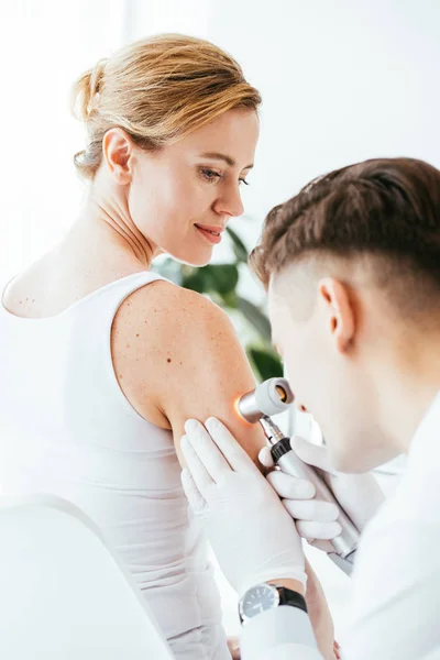 Dermatólogo en guantes de látex que sostiene dermatoscopio mientras examina hermoso paciente con enfermedad de la piel - foto de stock