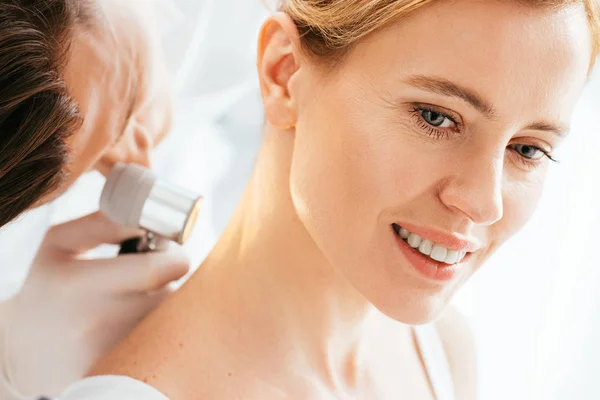 Selective focus of attractive woman smiling while dermatologist examining neck with dermatoscope — Stock Photo