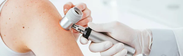 Prise de vue panoramique du dermatologue en gant de latex tenant le dermatoscope tout en examinant le patient — Photo de stock