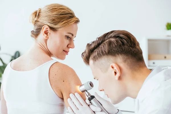 Dermatólogo en guantes de látex que sostiene dermatoscopio mientras examina hermosa mujer con enfermedad de la piel - foto de stock