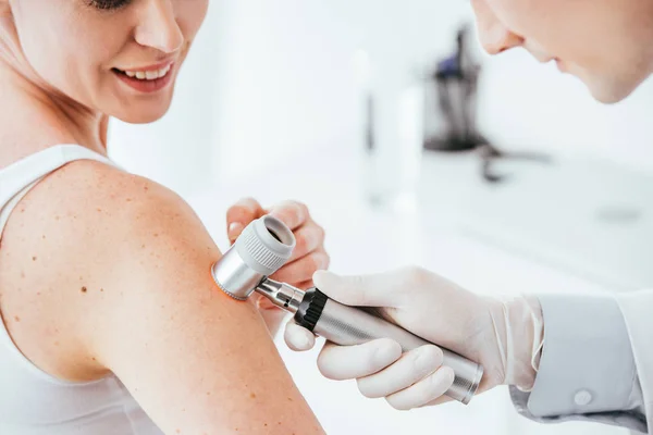 Visão cortada de dermatologista segurando dermatoscópio enquanto examina mulher alegre com doença de pele — Fotografia de Stock