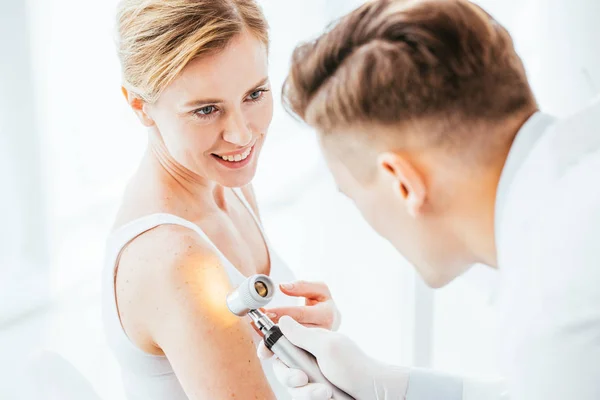 Selektiver Fokus der fröhlichen Frau, die den Dermatologen mit dem Dermatoskop anschaut, während sie die Hand untersucht — Stockfoto