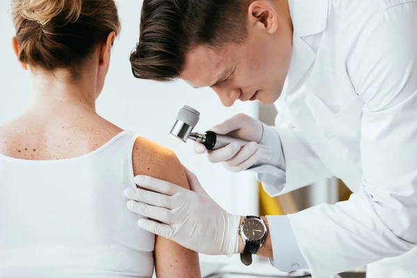 Dermatólogo guapo sosteniendo dermatoscopio mientras examina paciente - foto de stock