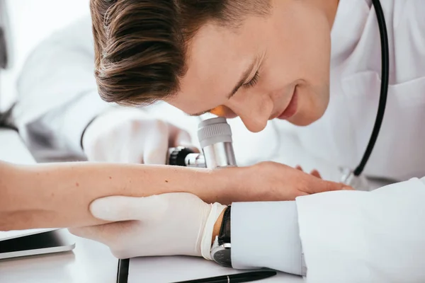 Cheerful dermatologist examining hand of woman while holding dermatoscope — Stock Photo