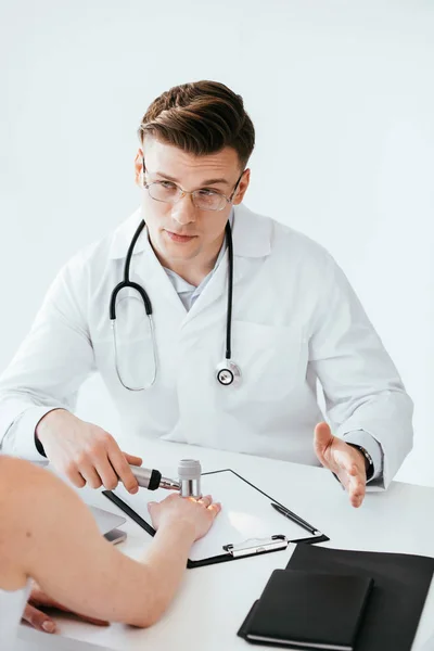 Foco seletivo de dermatologista bonito em óculos segurando dermatoscópio enquanto olha para o paciente — Fotografia de Stock