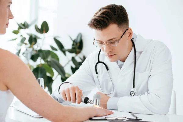 Selective focus of dermatologist in glasses examining woman while holding dermatoscope in clinic — Stock Photo