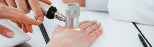 Panoramic shot of dermatologist examining hand of woman while holding dermatoscope in clinic — Stock Photo