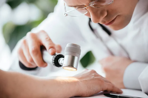 Selektiver Fokus des Dermatologen in der Brille, die das Dermatoskop in der Nähe der Hand des Patienten hält — Stockfoto