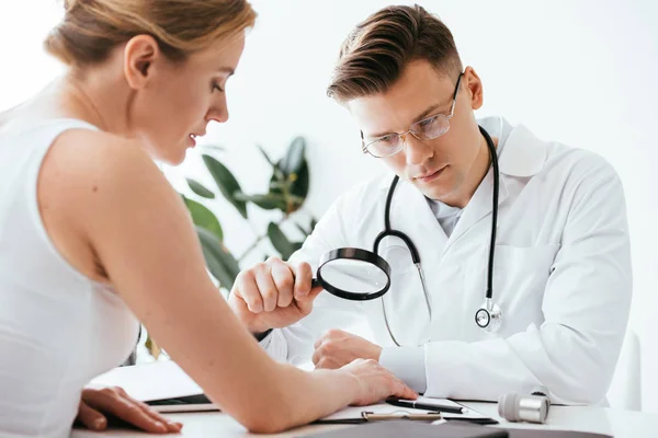 Selective focus of handsome dermatologist in glasses holding magnifying glass while examining woman in clinic — Stock Photo