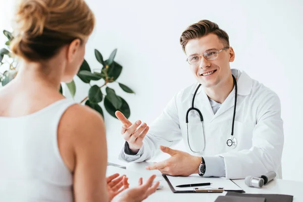 Messa a fuoco selettiva del medico allegro in occhiali e cappotto bianco gesticolando mentre si guarda paziente — Foto stock