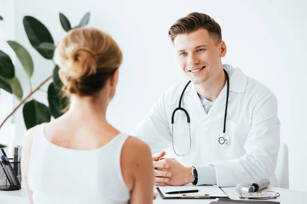 Вибірковий акцент веселого лікаря в білому пальто сидить з затиснутим рук і дивиться на пацієнта — стокове фото