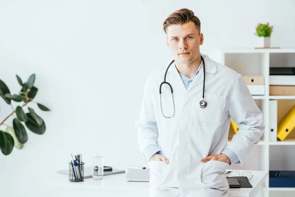 Guapo médico de bata blanca de pie con las manos en los bolsillos en la clínica - foto de stock