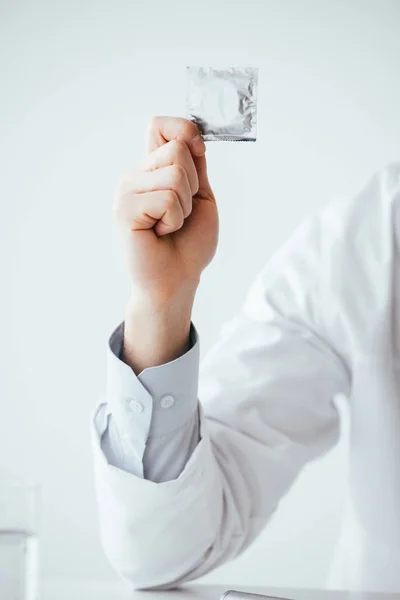 Vista recortada del médico de bata blanca sosteniendo el condón en la mano - foto de stock