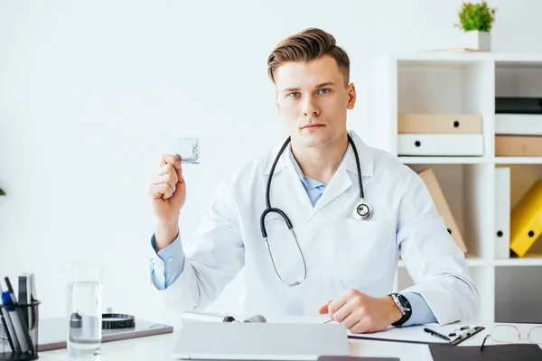 Handsome and serious doctor in white coat holding condom in hand — Stock Photo