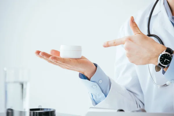 Cropped view of doctor in white coat pointing with finger at container with cosmetic cream — Stock Photo