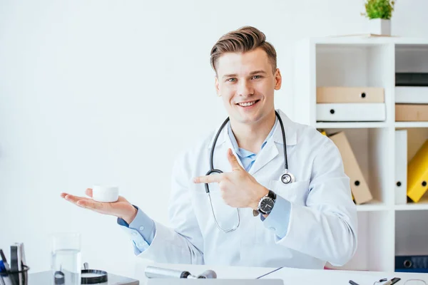 Gutaussehender und fröhlicher Dermatologe zeigt mit dem Finger auf kosmetische Creme in der Klinik — Stockfoto