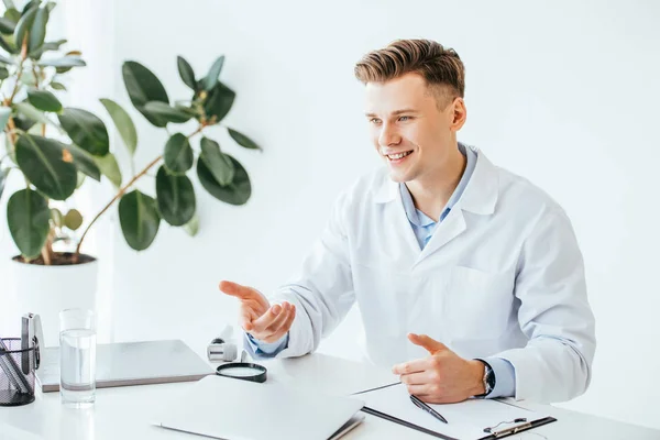 Красивый и счастливый доктор в белом халате улыбается во время жеста в клинике — стоковое фото